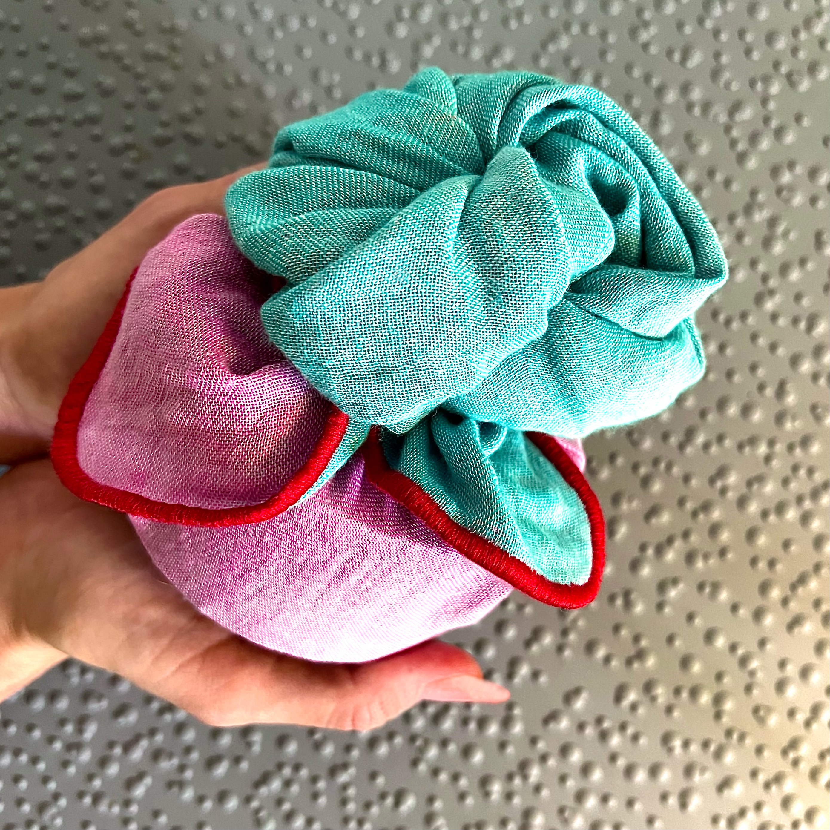 Gift artfully wrapped to resemble a rose using the 'Sparkling Reflections' reusable fabric gift wrap from COVER, showcasing vibrant shades of pink and turquoise in a flower fold. The beautifully wrapped gift is delicately held by two hands against a muted grey background.